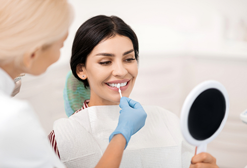 Dentist holding a veneer in front of a smiling patient's teeth as she looks in a mirror