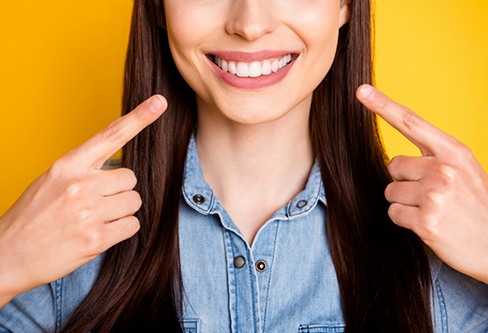 Brunette woman with straight white teeth pointing to her smile